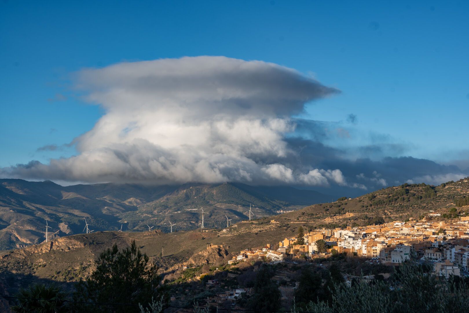 Giant lenticular stacked clouds above some hillsides with a small spanish town highlighted in the sunlight