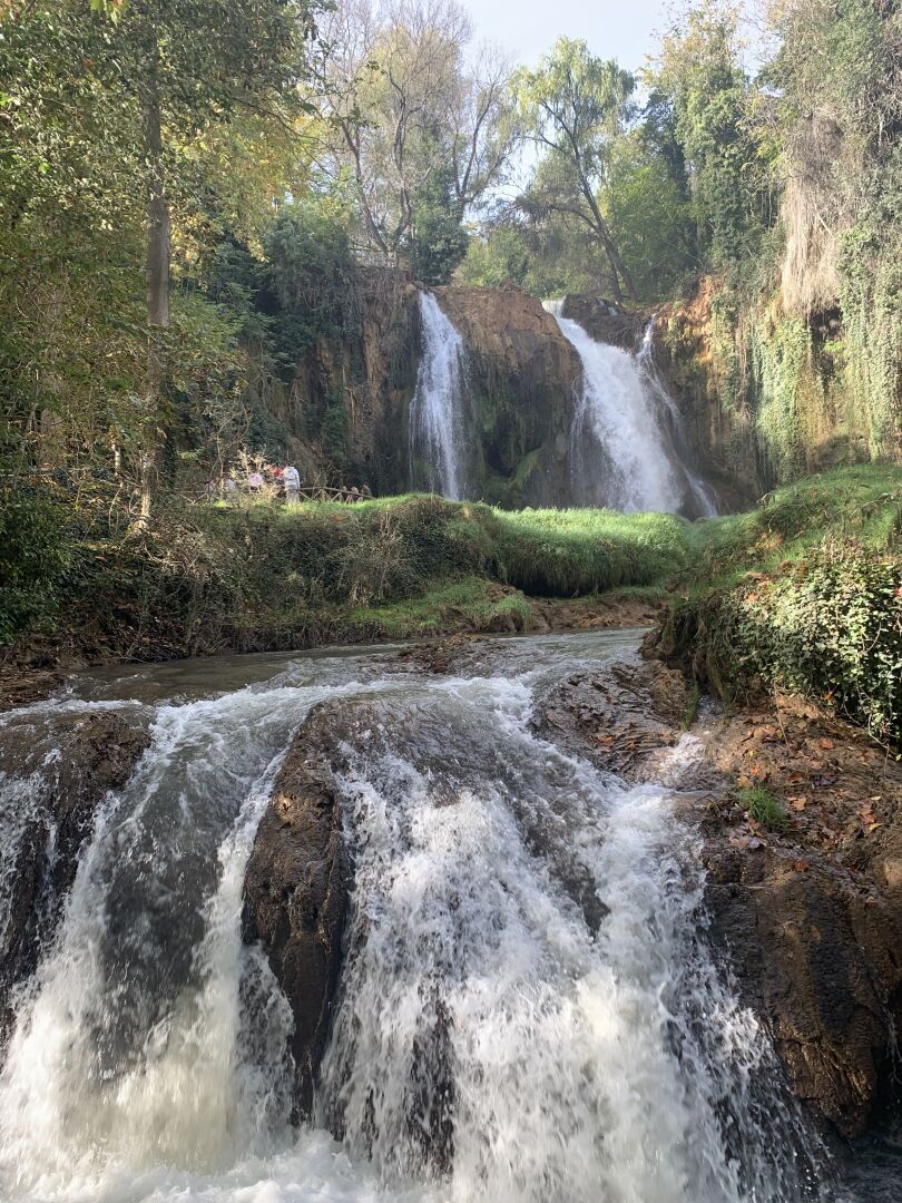 Two cascades of waterfalls flow through an autumn forest setting on a sunny day.