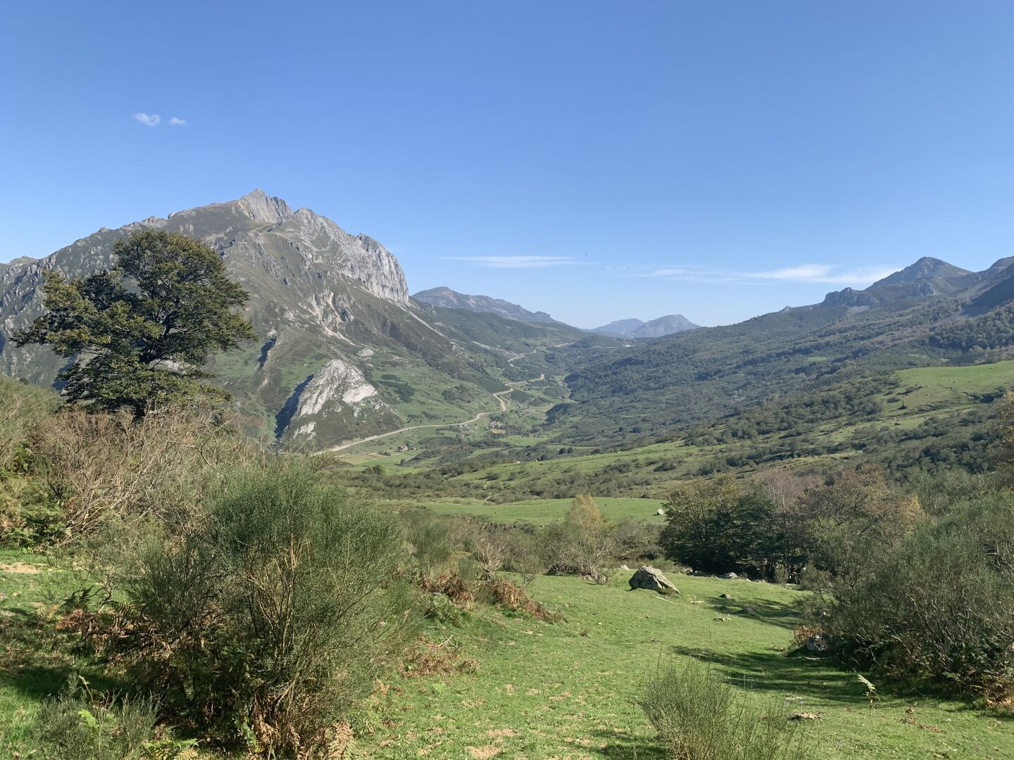 A green mountain setting with taller rockier mountains in the background on a very sunny autumn day.