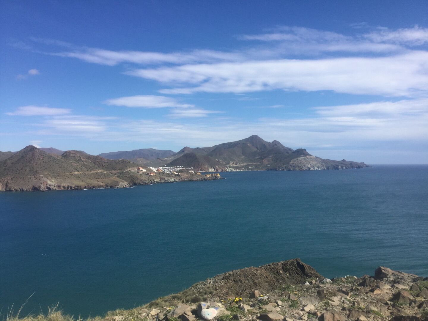 A coastal view featuring rugged mountains and cliffs, with a calm blue sea in the foreground. The sky is partly cloudy, and a small town is visible along the shore.