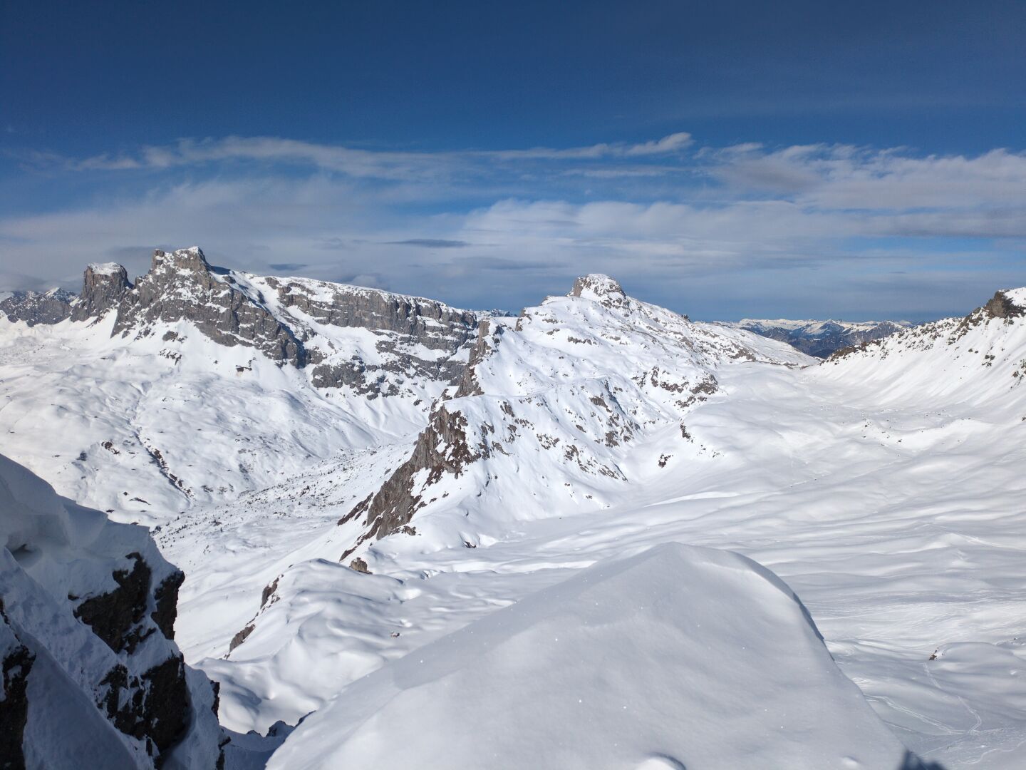 Snow covered mountains and a mostly blue sky with some thin white clouds in the foreground. The clouds are darker the farther away. In the center the snow covered plateau of Plassegga. On its right the cummits of Graustei, Mittelflua and Schijenflue.  At the left the rocky face of the Sulzflue.